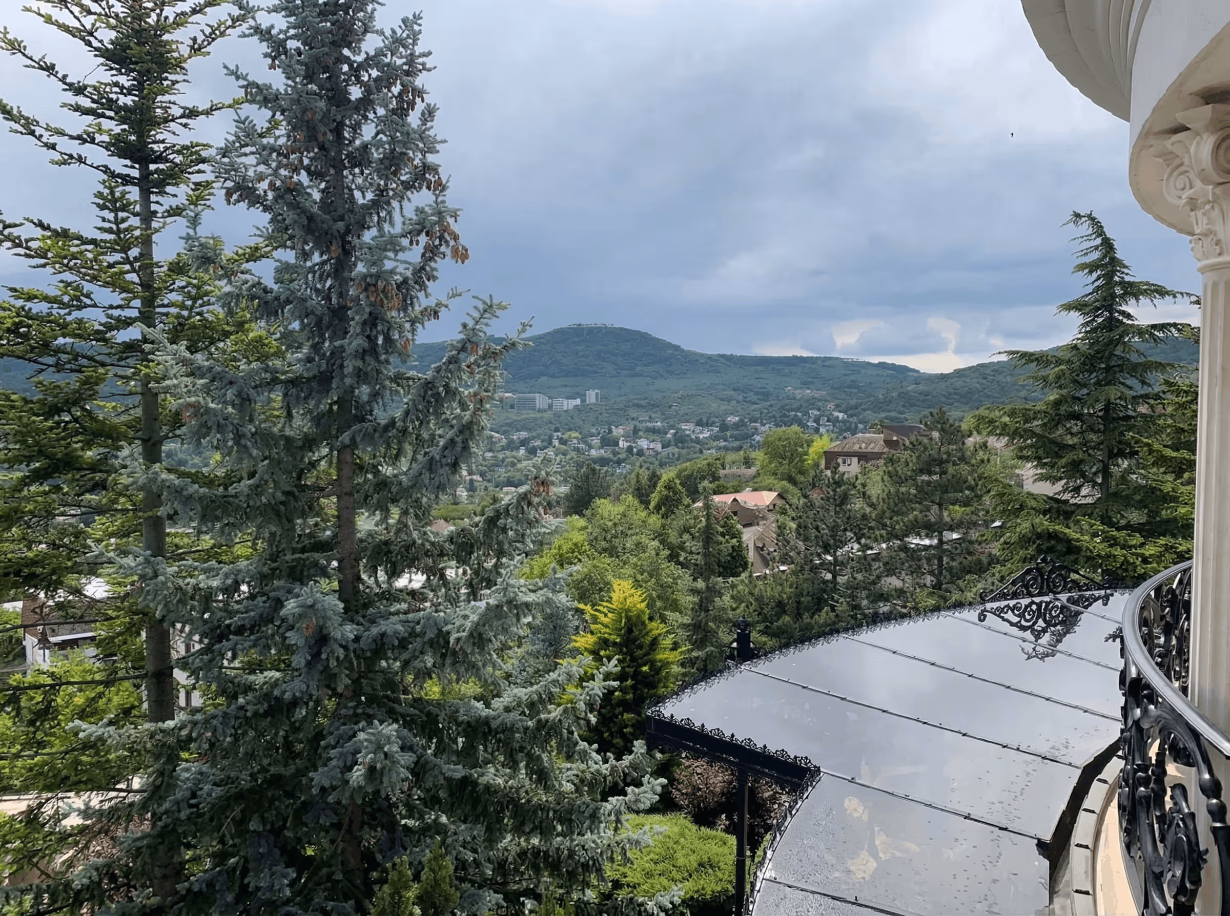 View of green trees and hills under a cloudy sky from a balcony with ornate railing.