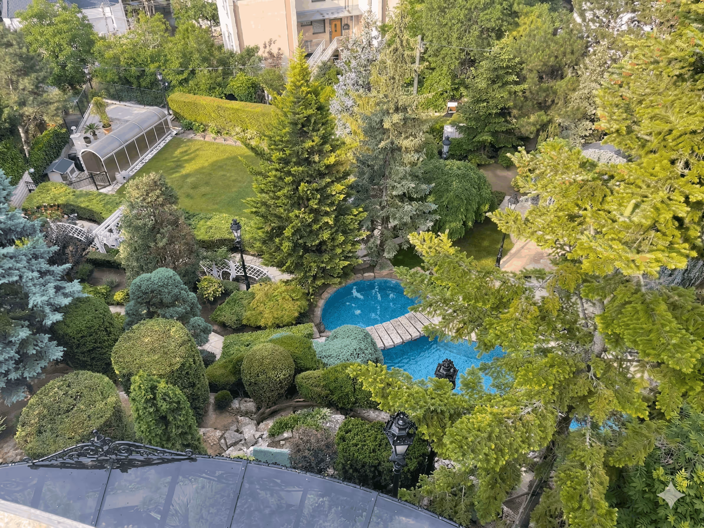 Aerial view of a lush garden with trimmed bushes, tall trees, a blue pond, and stone pathway bridge.