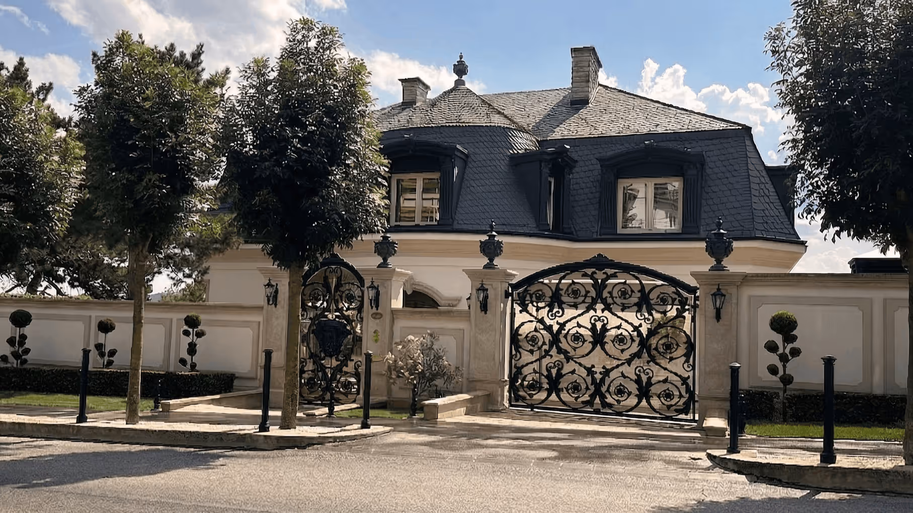 Elegant mansion with ornate black wrought iron gates, trimmed trees, and blue sky with clouds.