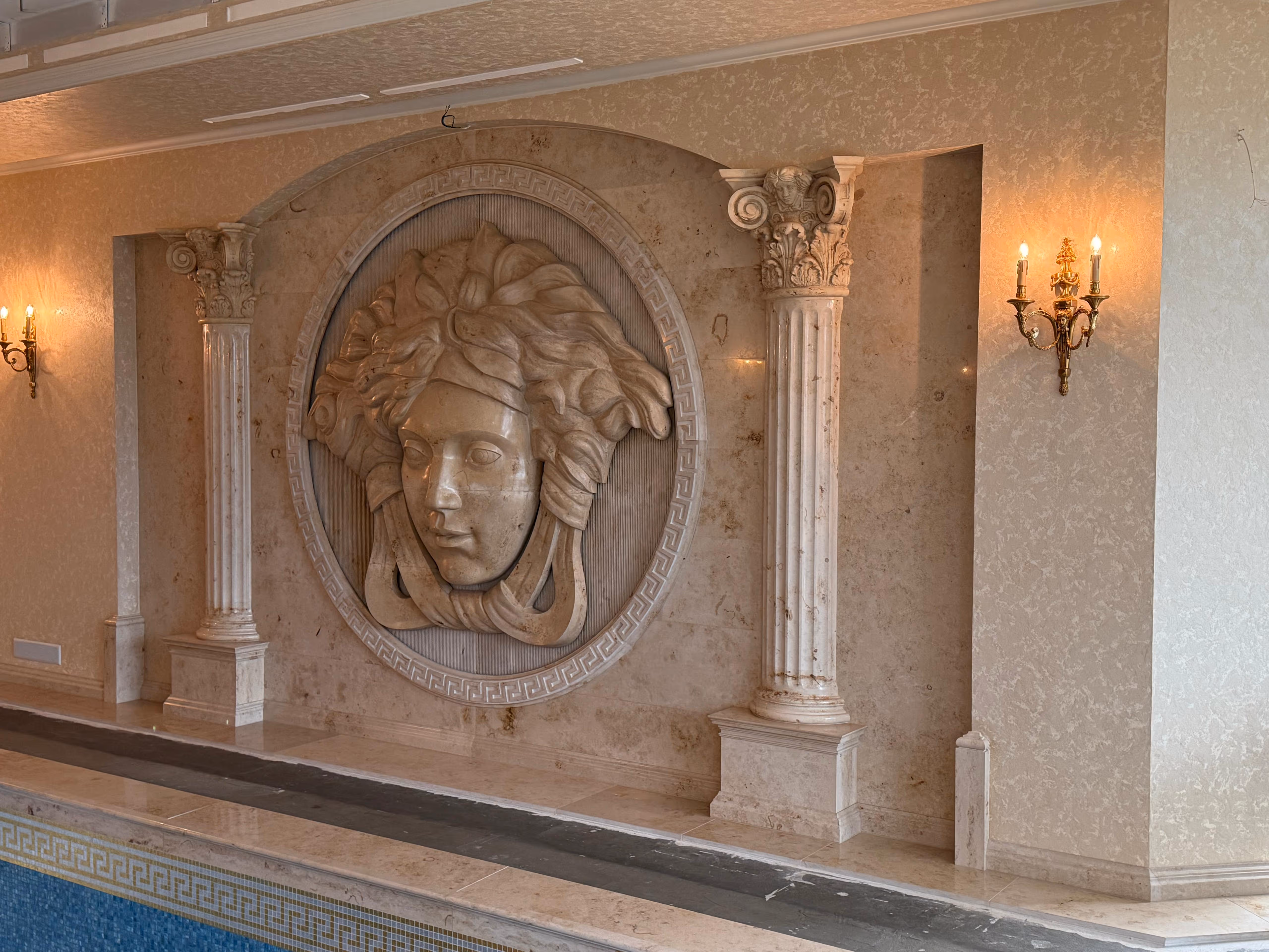 Interior wall with classical columns and a large carved medallion of a face between two lit wall sconces.