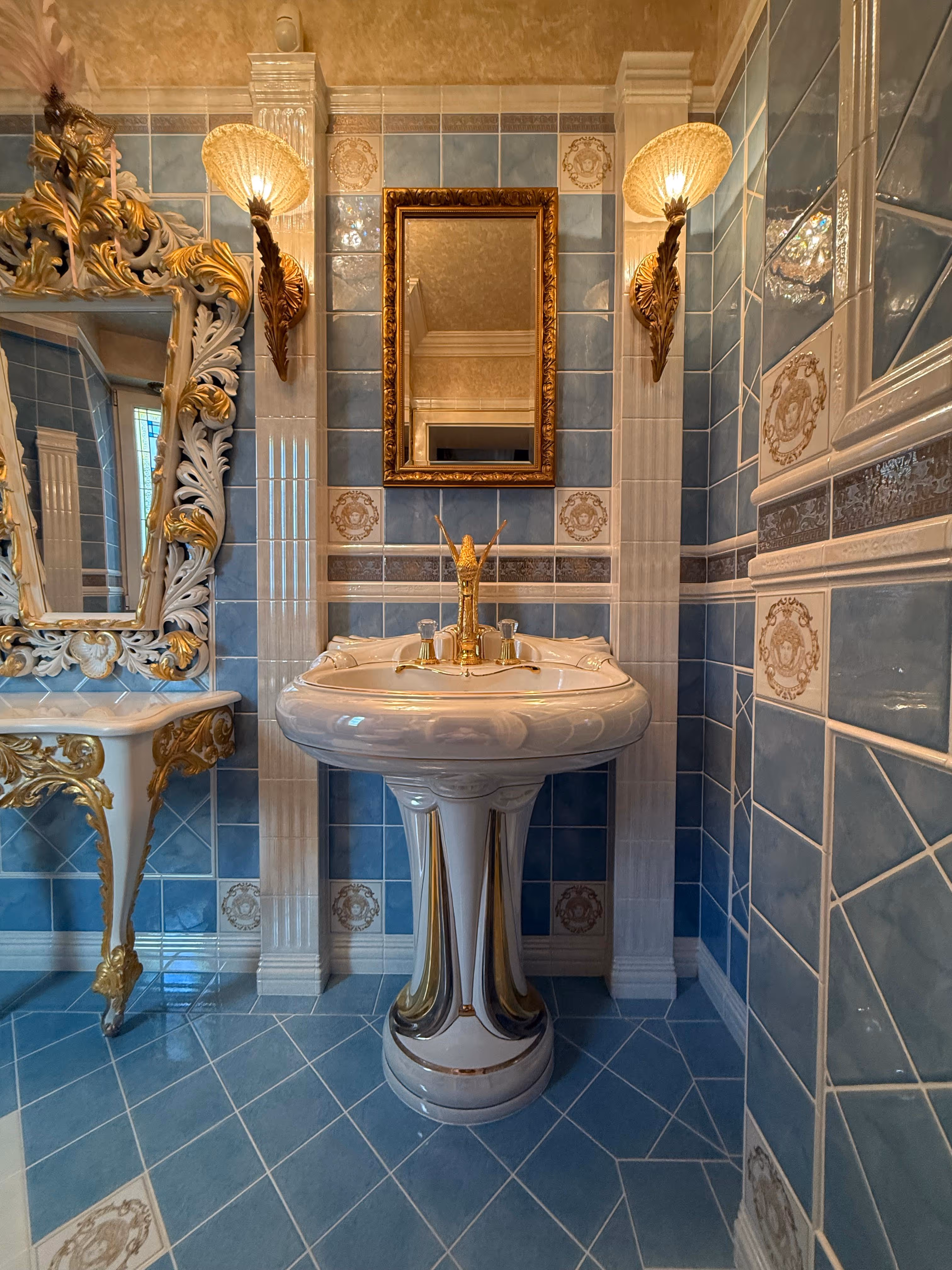 Ornate vintage bathroom sink with gold fixtures, framed mirror, decorative lamps, and blue tiled walls.