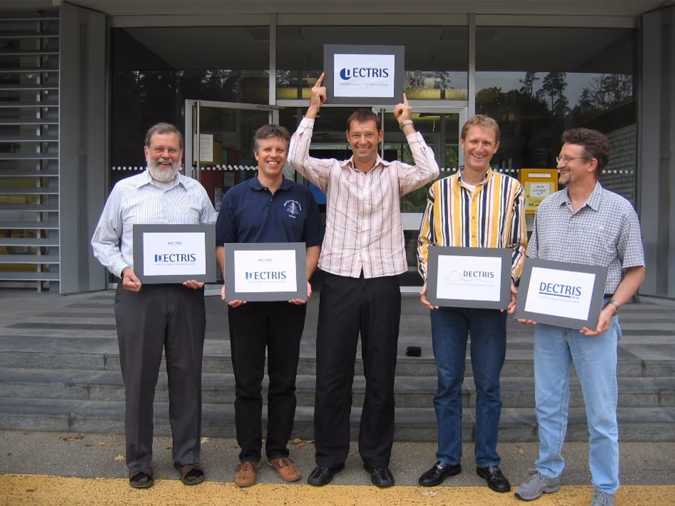 Five men standing outside a glass entrance holding signs with the word 'DECTRIS' in blue letters, one man in the center pointing upwards to another DECTRIS sign above.