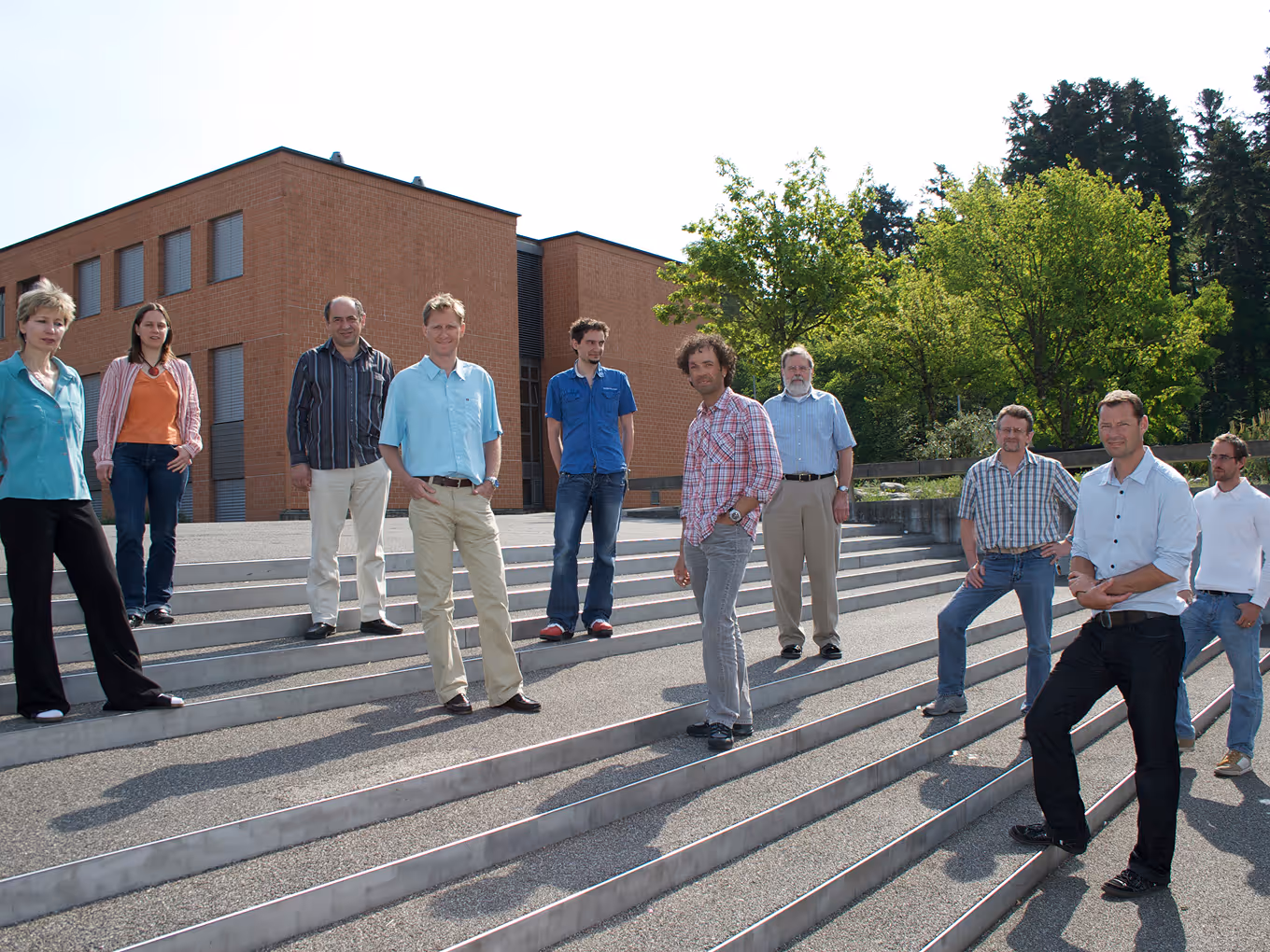 Group of nine adults standing on outdoor steps in front of a brick building and green trees, posing for a photo.