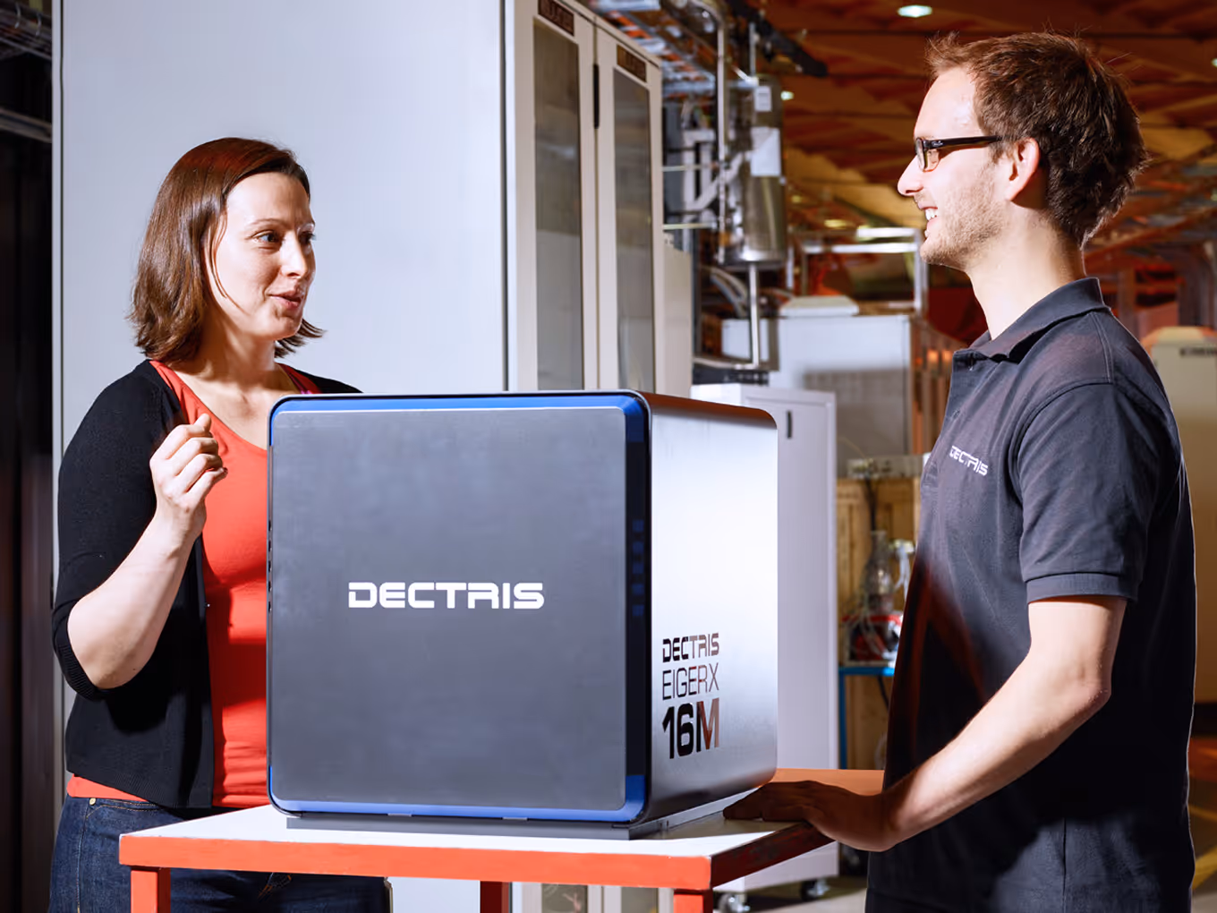 Two people standing and talking next to a Dectris Eiger X 16M scientific detector device on a table in a lab setting.