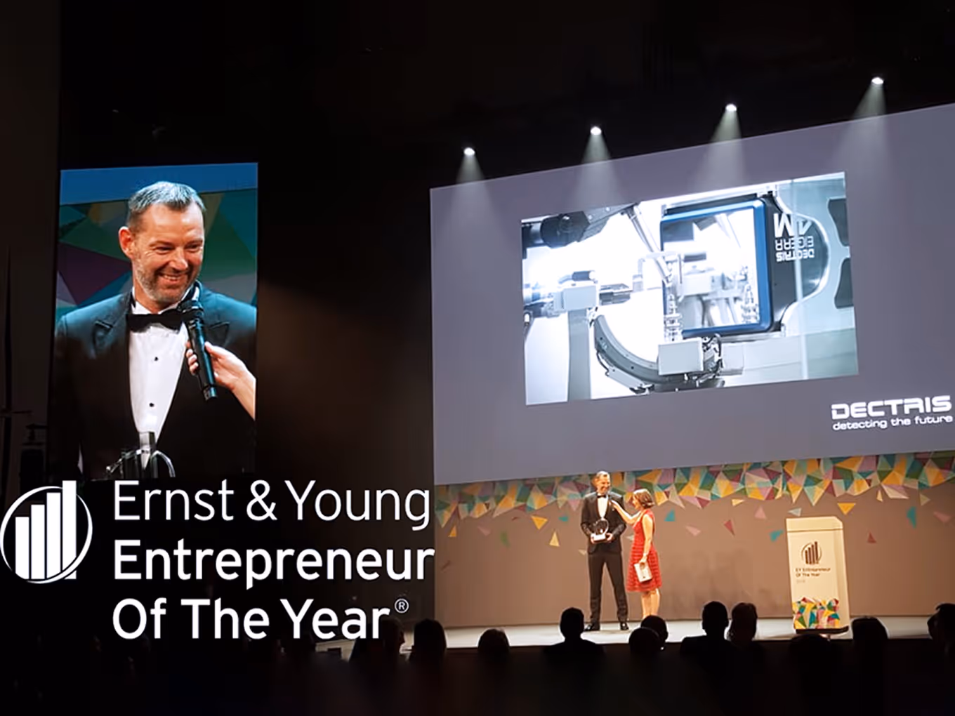 Man in tuxedo receiving an award on stage with a woman in a red dress holding a microphone at the Ernst & Young Entrepreneur Of The Year event.