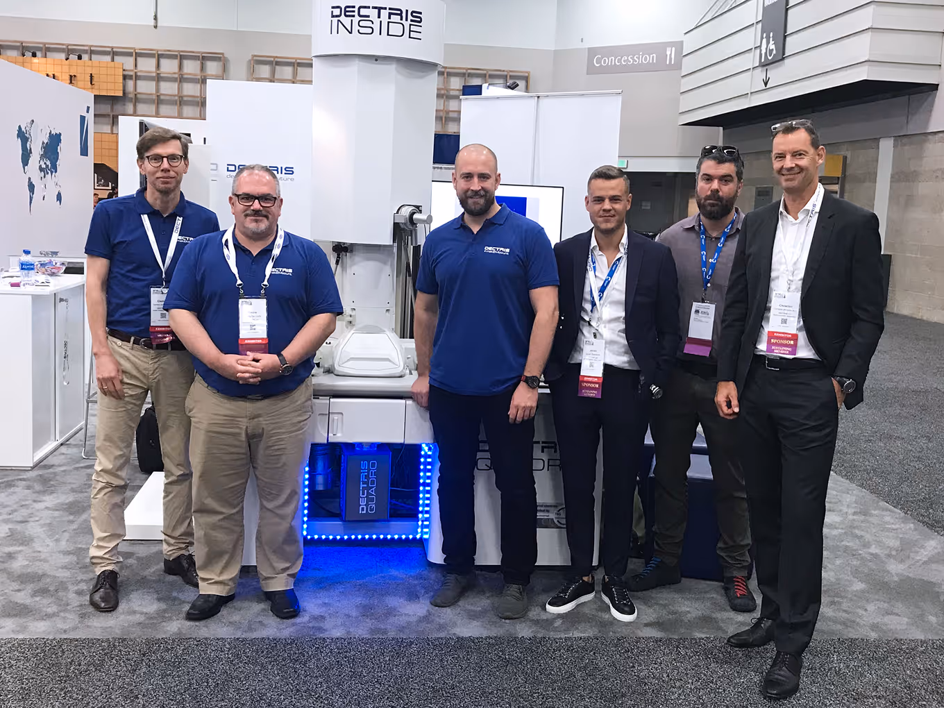 Six men standing in front of a Dectris Inside exhibition booth with scientific equipment, some wearing blue Dectris polo shirts and conference badges.
