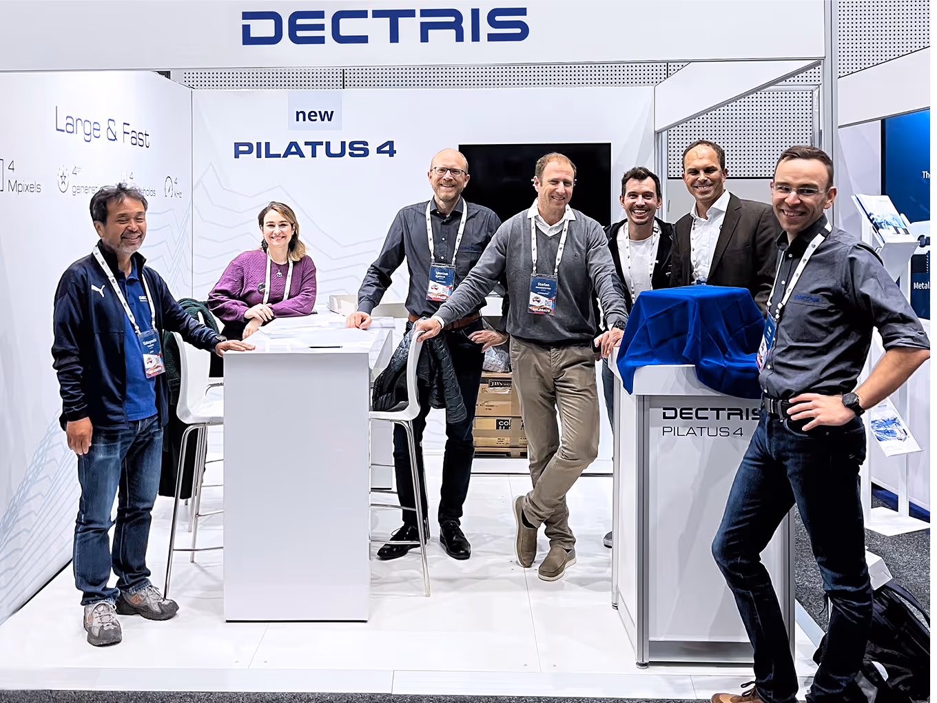 Seven people posing and smiling at a DECTRIS exhibition booth featuring the PILATUS 4 product.