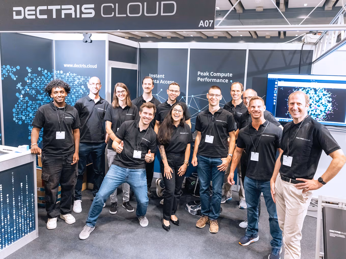 Group of eleven people wearing black Dectris Cloud polo shirts posing and smiling in a tech booth with digital graphics in the background.