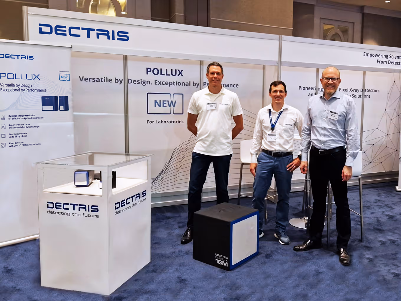 Three men standing in a booth with Dectris branding and promotional displays for Pollux X-ray detectors at an exhibition.