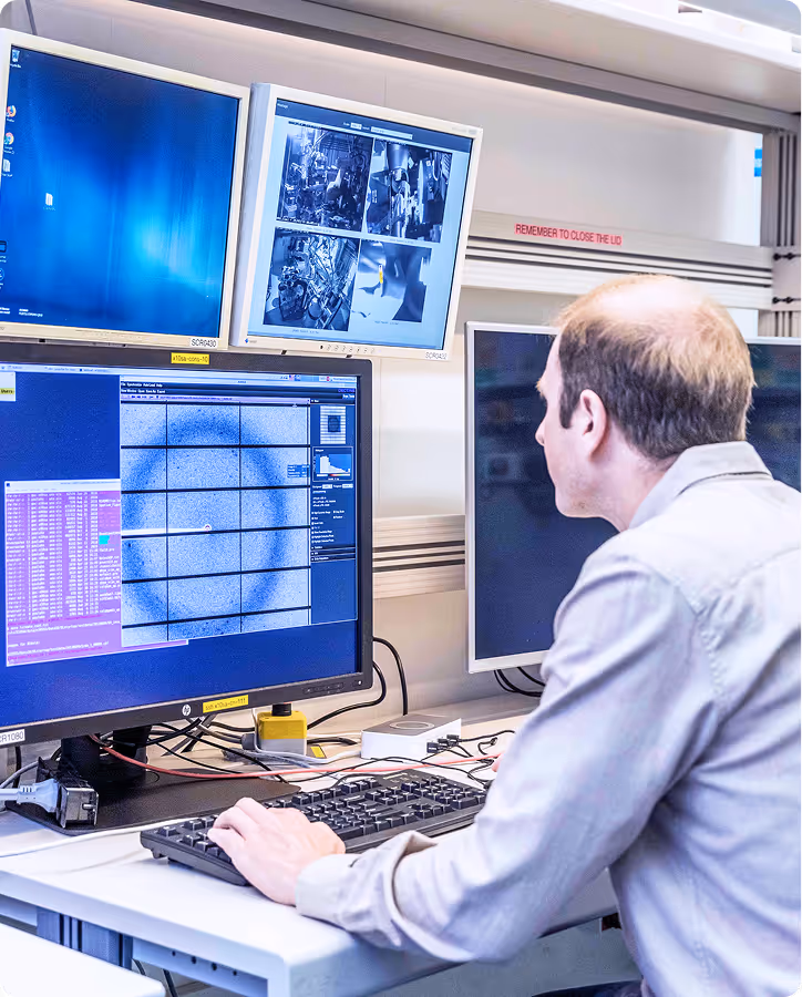 Man working at a desk with multiple monitors displaying technical and scientific data in a lab setting.