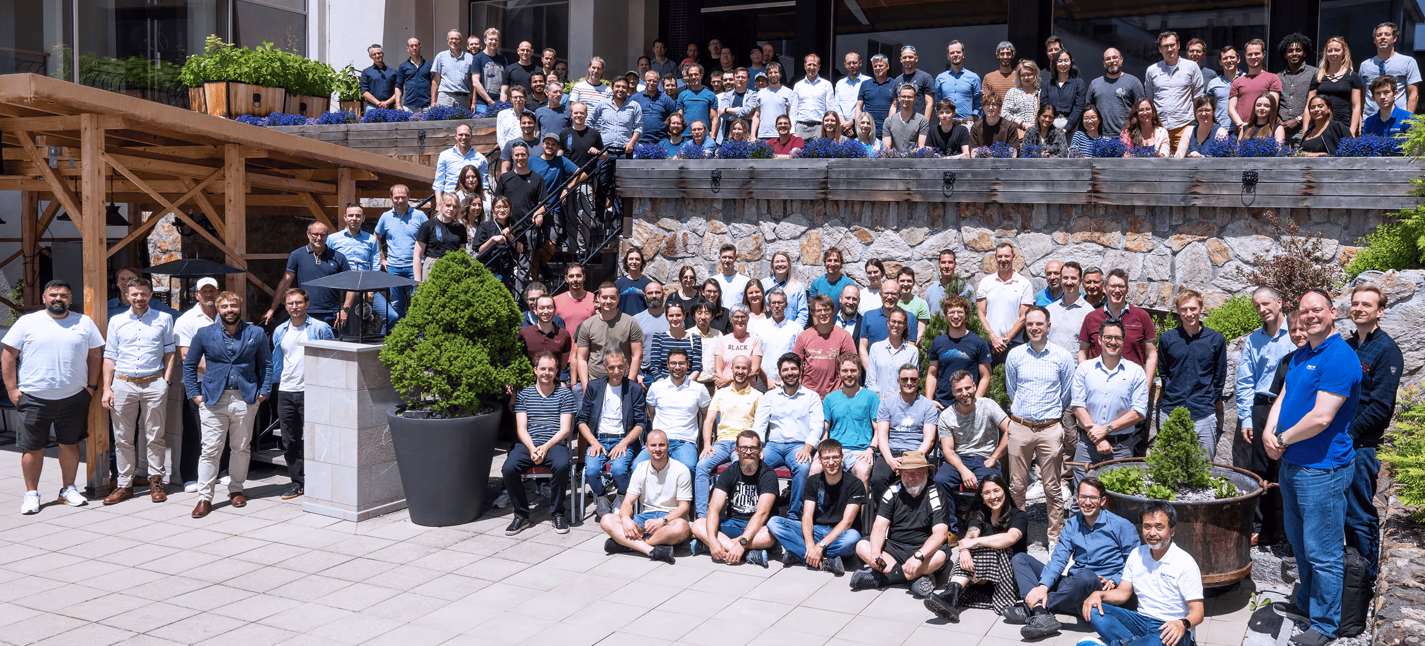 Large group of people posing outdoors in front of a building with stone walls and wooden beams, under bright sunlight.