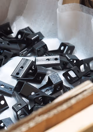 A pile of small, black metal brackets with holes, placed on a white surface inside a box.