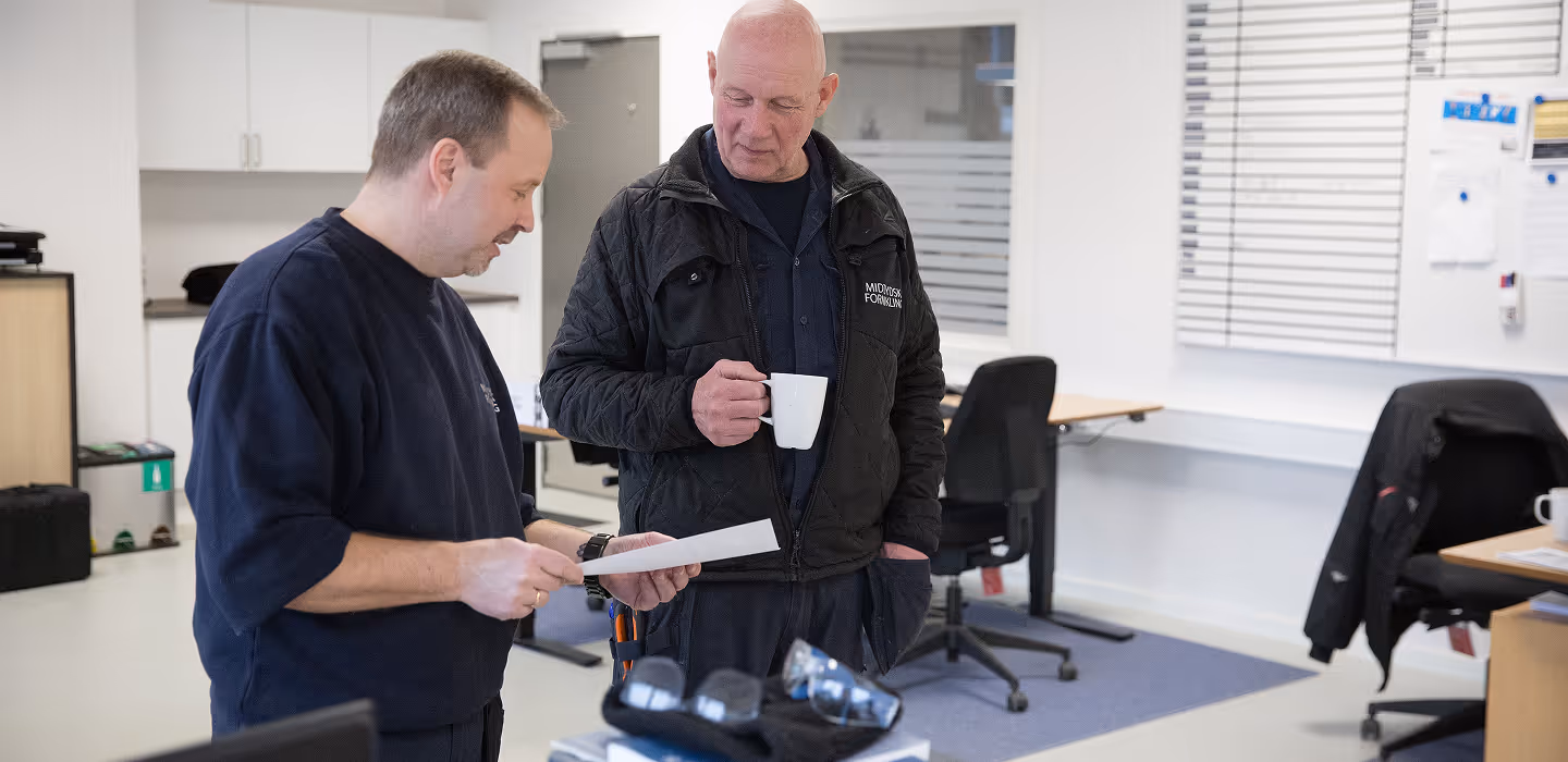 Two men in an office discussing a document while one holds a white coffee mug.