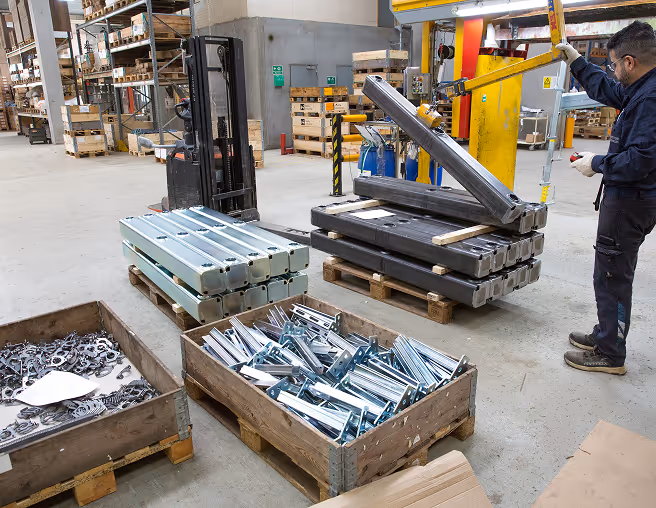 Worker operating a machine lifting metal parts in a warehouse with stacked metal items on pallets.