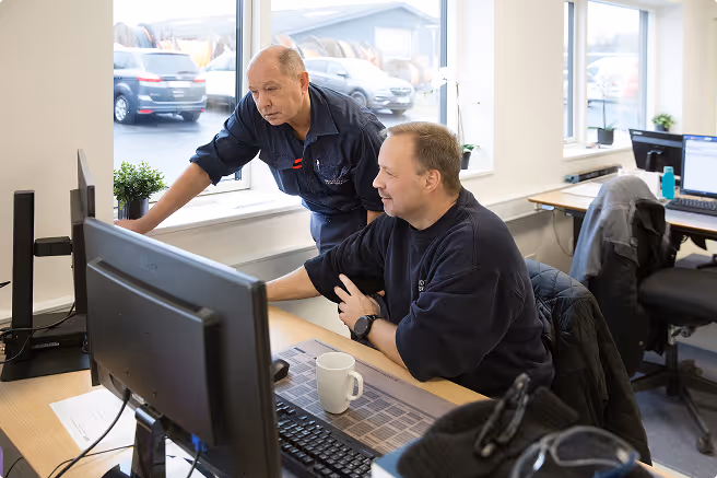Two men working together at a desk with computers in a bright office with large windows.