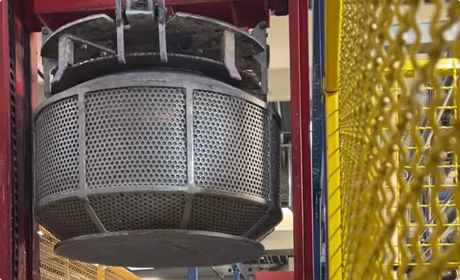Large industrial metal basket with perforations suspended in a factory setting near yellow safety barriers.