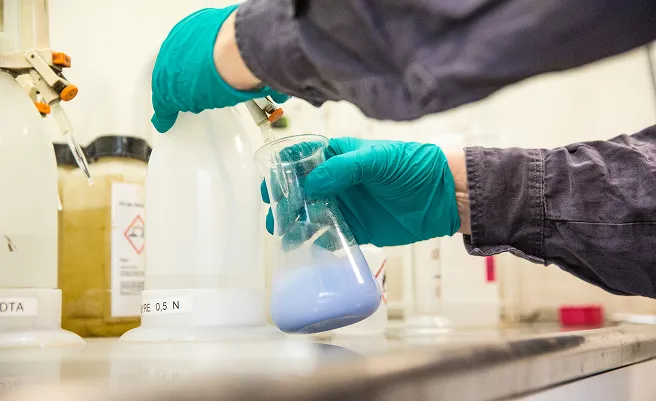 Hands in green gloves holding a flask with a blue chemical inside in a laboratory.