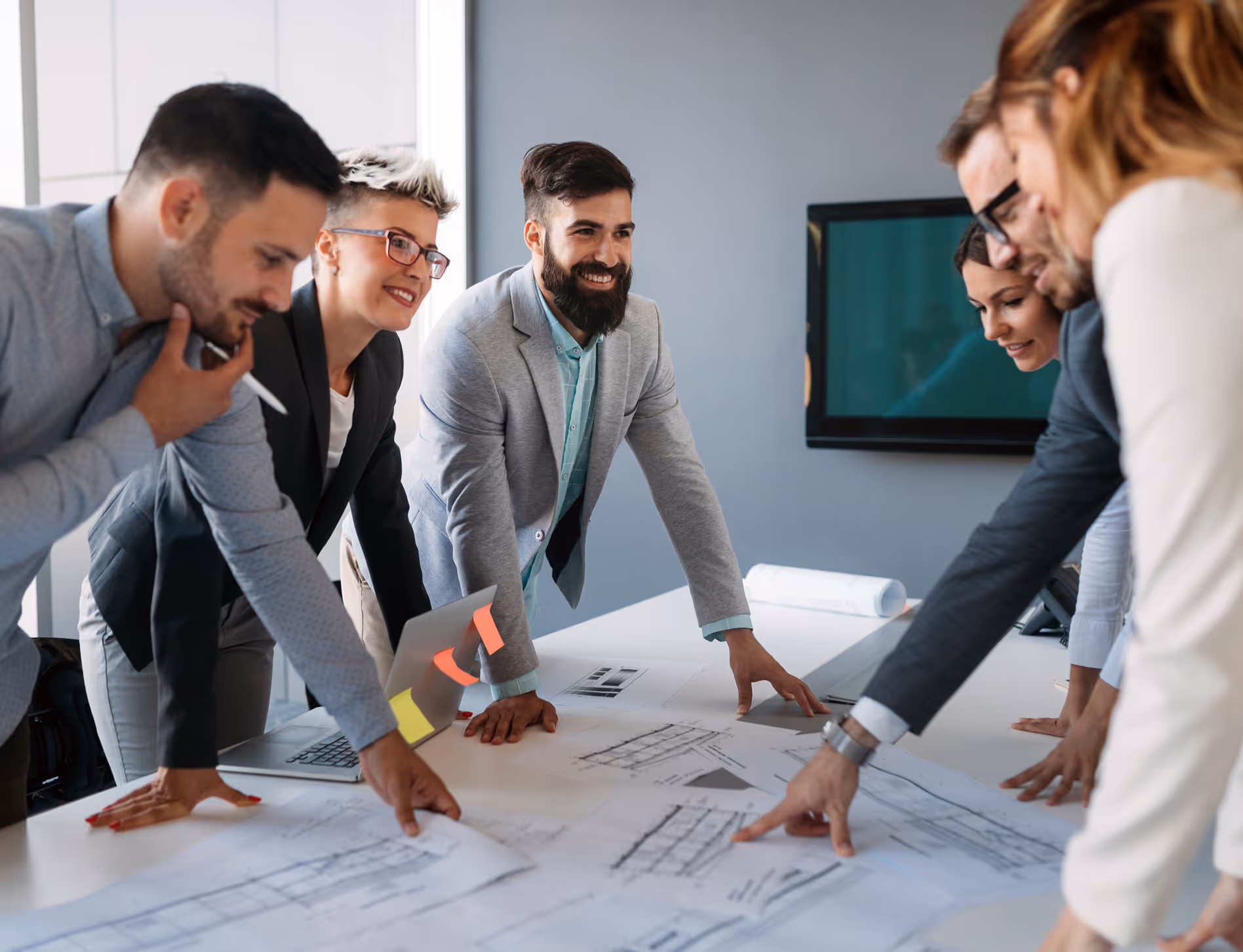 Group of six professionals smiling and discussing architectural plans on a table in an office.