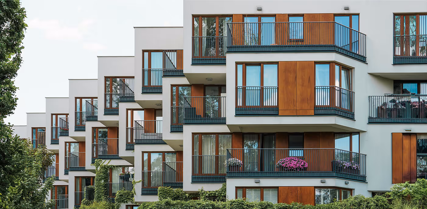 Modern white apartment building with brown wood panels, balconies, and greenery in front.
