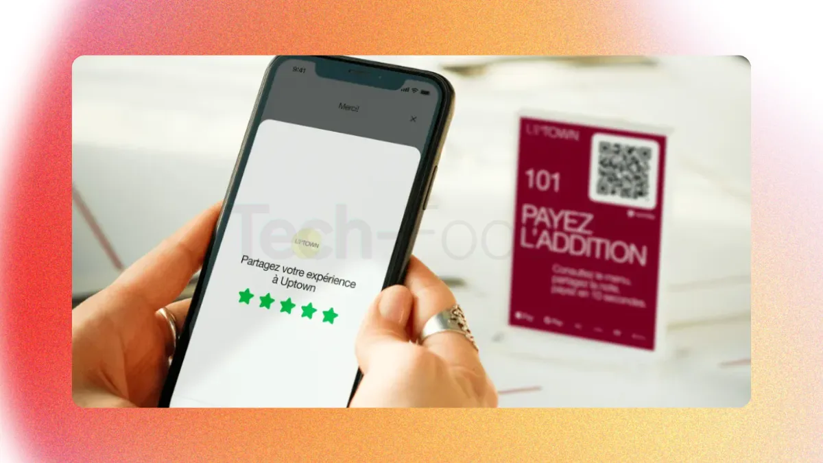 Gros plan sur les mains d'un client tenant son smartphone au-dessus d'une table de restaurant. L'écran affiche l'interface de dépôt d'avis Google avec 5 étoiles cochées. En arrière-plan flou, on distingue le chevalet avec le QR Code de l'établissement.