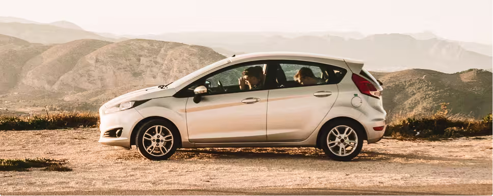 Silver compact car parked on a dirt road with two people inside and mountains in the background.
