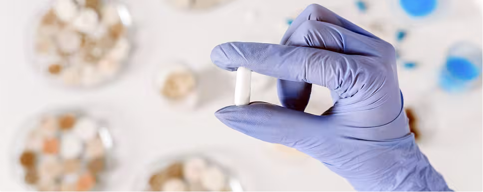 Gloved hand holding a single white pill with blurred pills in containers in the background.