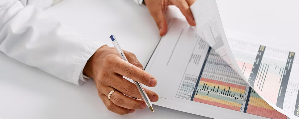 Person in white coat holding a pen and reviewing a printout with colored charts and data tables.
