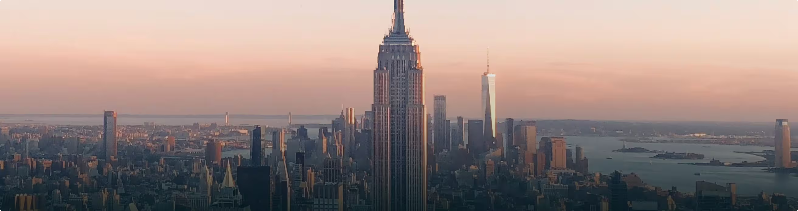 City skyline at sunset with Empire State Building prominently in center and One World Trade Tower in background.