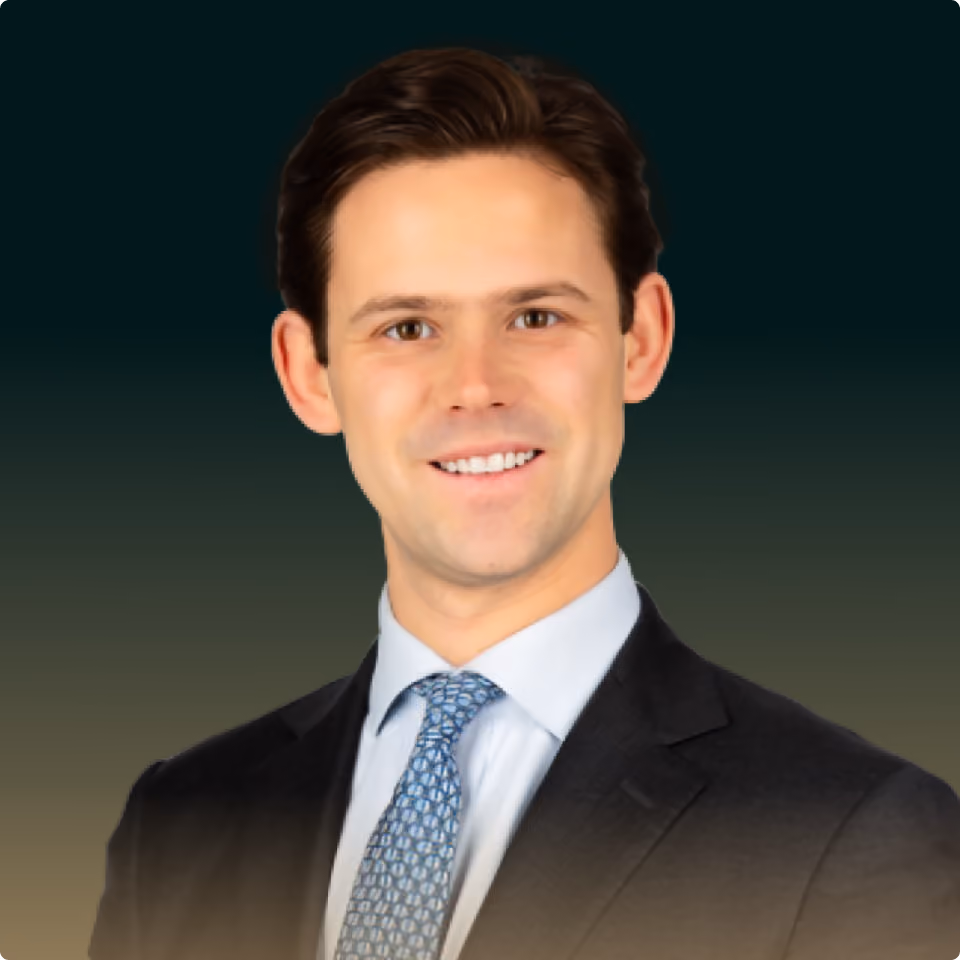 Smiling young man with brown hair wearing a black suit, light blue shirt, and patterned tie.