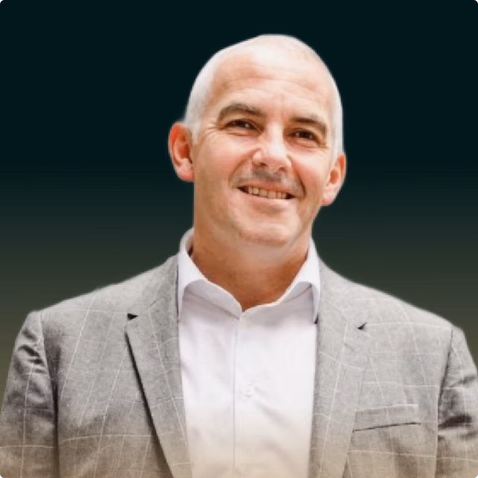 Smiling middle-aged man wearing a gray checkered suit jacket and white shirt against a dark background.