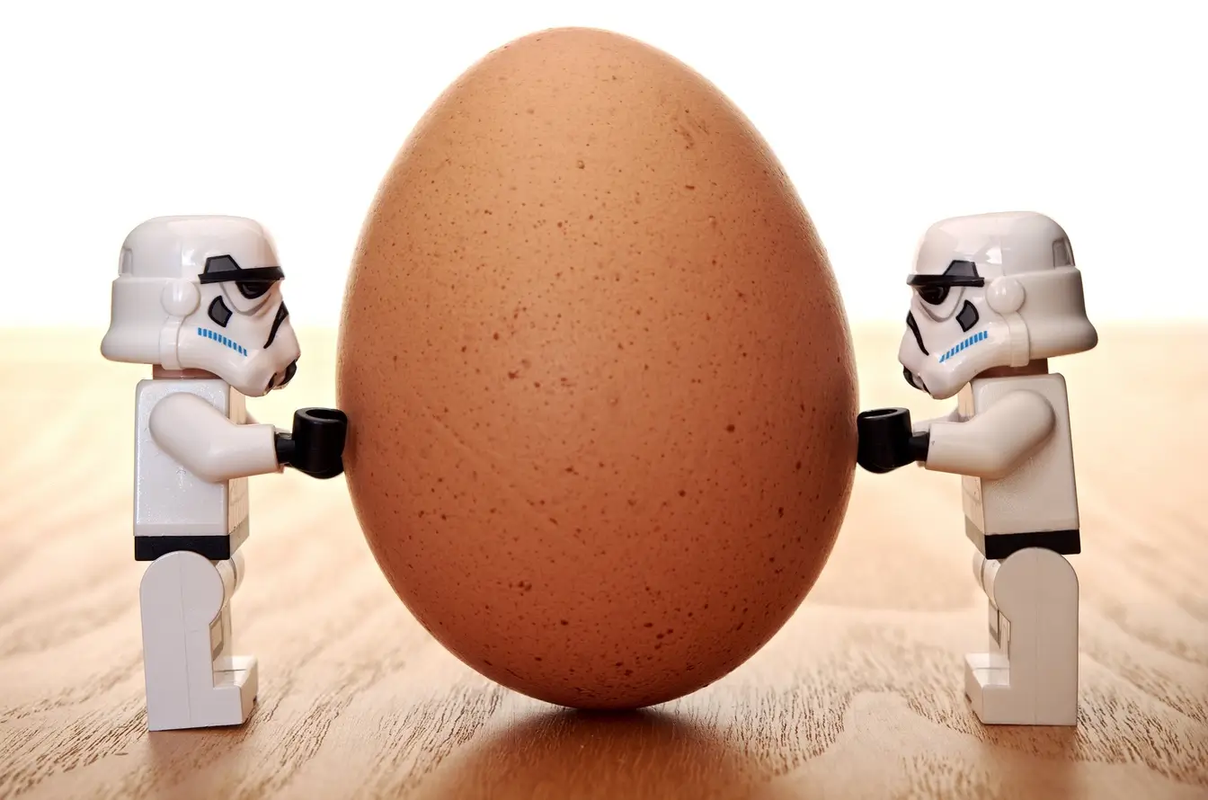 Almost identical toy astronauts standing on either side of an egg to represent the similarities and differences between procurement vs. purchasing.