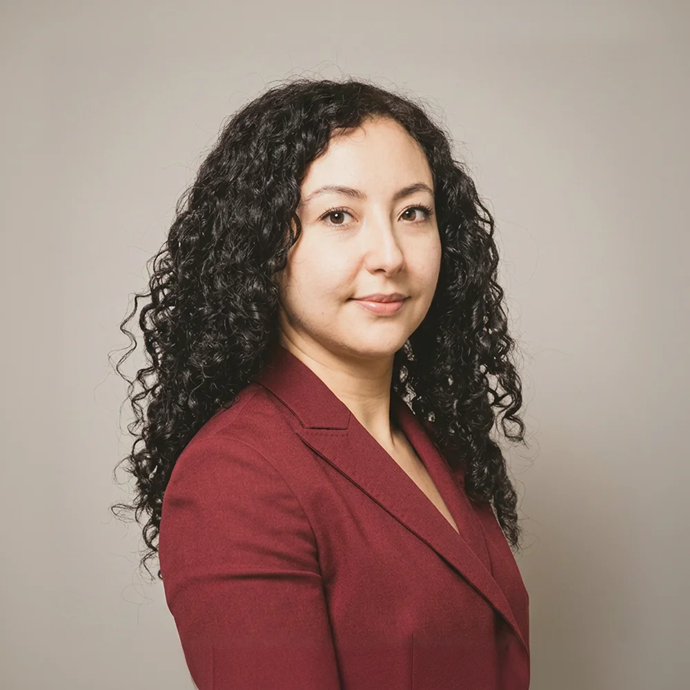 Portrait d'une femme aux cheveux noirs bouclés portant un blazer bordeaux, posant devant un fond gris uni.