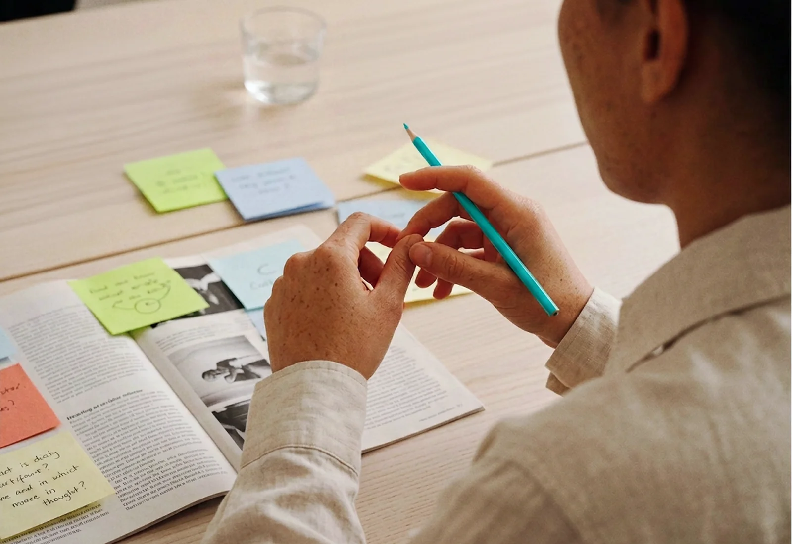 Person holding a turquoise pencil over a table with an open magazine and colorful sticky notes, with a glass of water in the background.