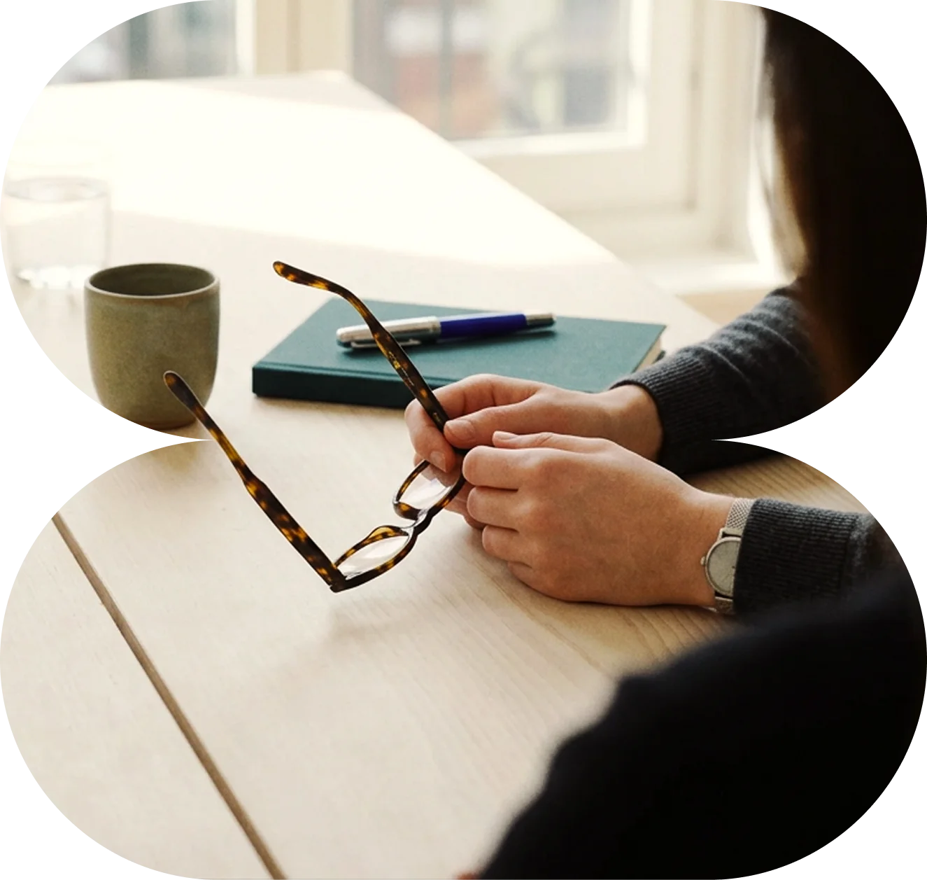 Person holding tortoiseshell glasses sitting at a desk with a green notebook, pen, and cup.