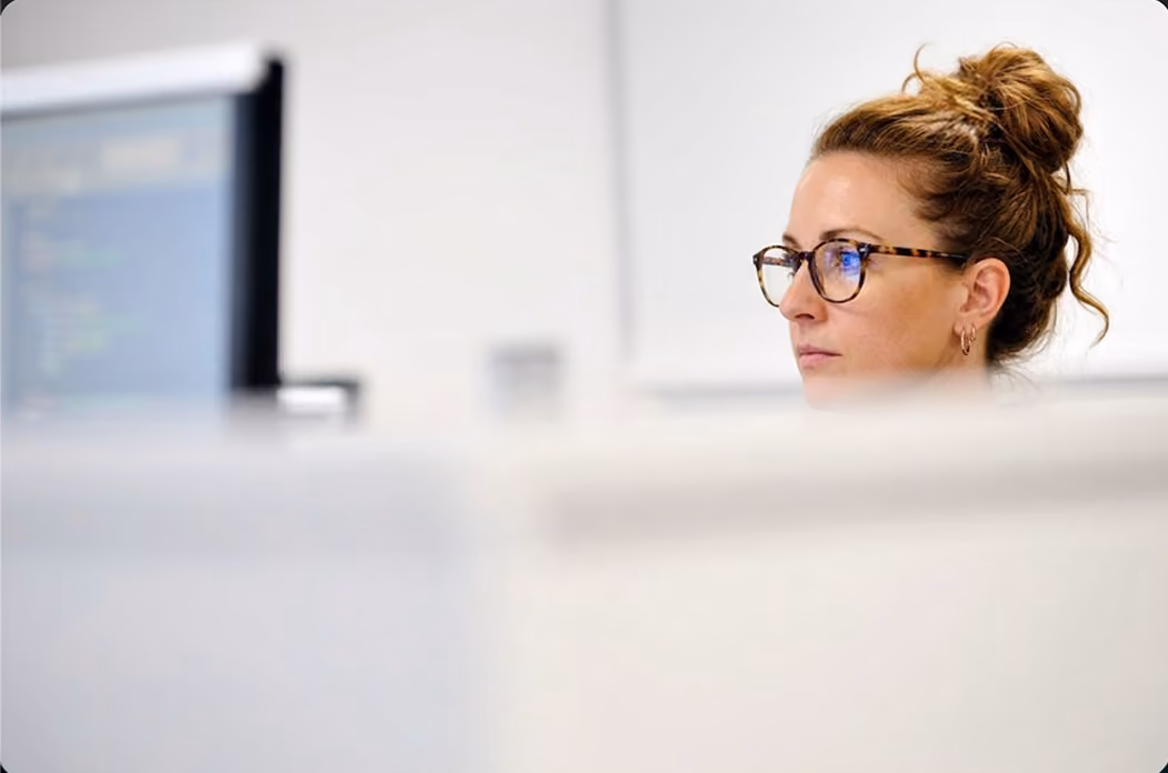 Femme aux cheveux attachés en chignon et portant des lunettes attentivement concentrée devant un écran d'ordinateur dans un bureau clair.