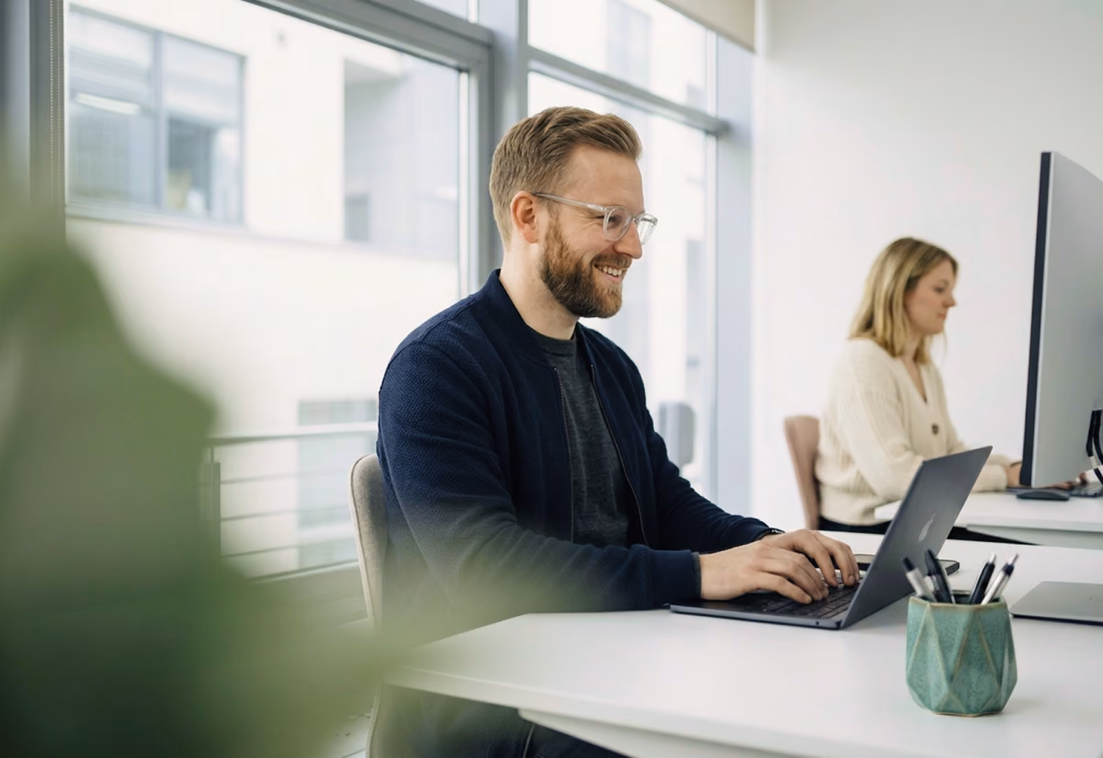 Un homme souriant avec des lunettes tapant sur un ordinateur portable dans un bureau lumineux, avec une femme travaillant sur un ordinateur en arrière-plan.