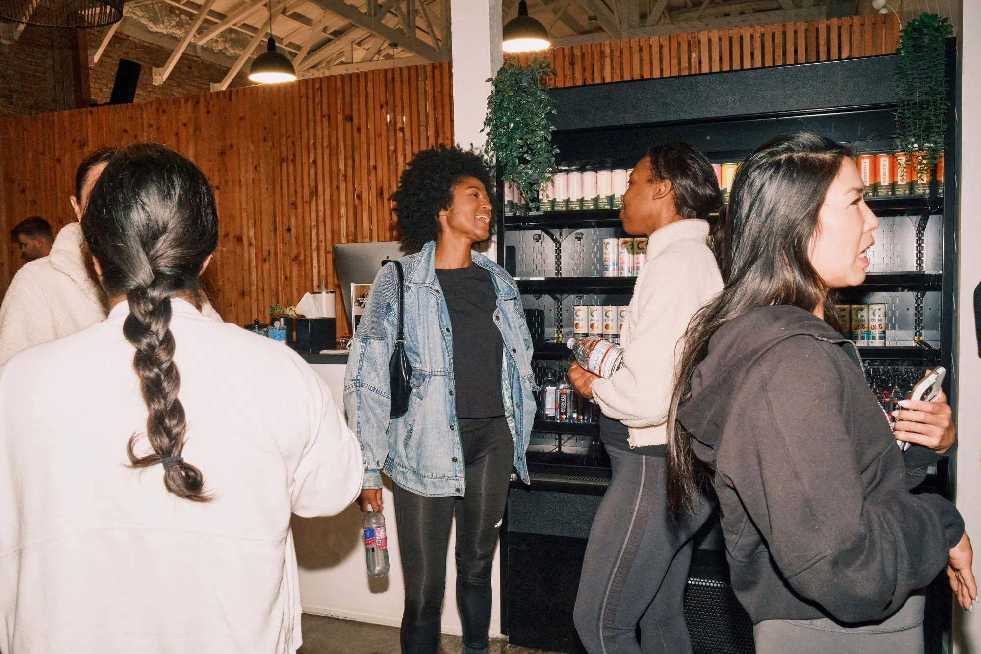 A group of women in comfortable athletic and casual clothing chat and smile together while holding water bottles. They are standing near a stocked beverage refrigerator in a modern lounge area featuring wood-slat walls.