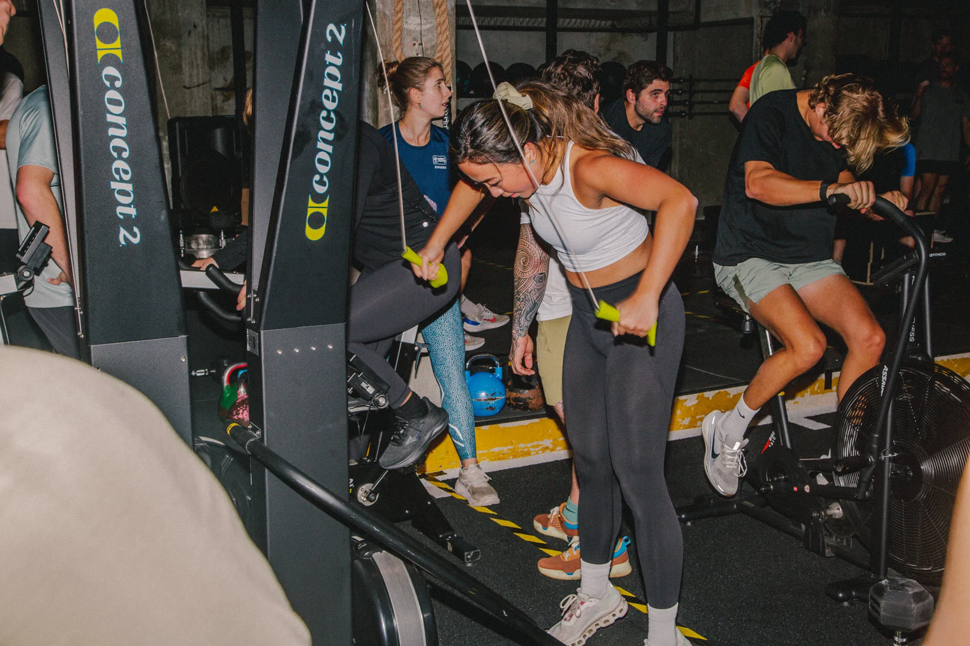 Several people are engaged in a high-intensity interval training class in a dimly lit gym. In the foreground, a woman actively pulls down the handles of a Ski machine, while a man next to her pedals hard on an air bike.
