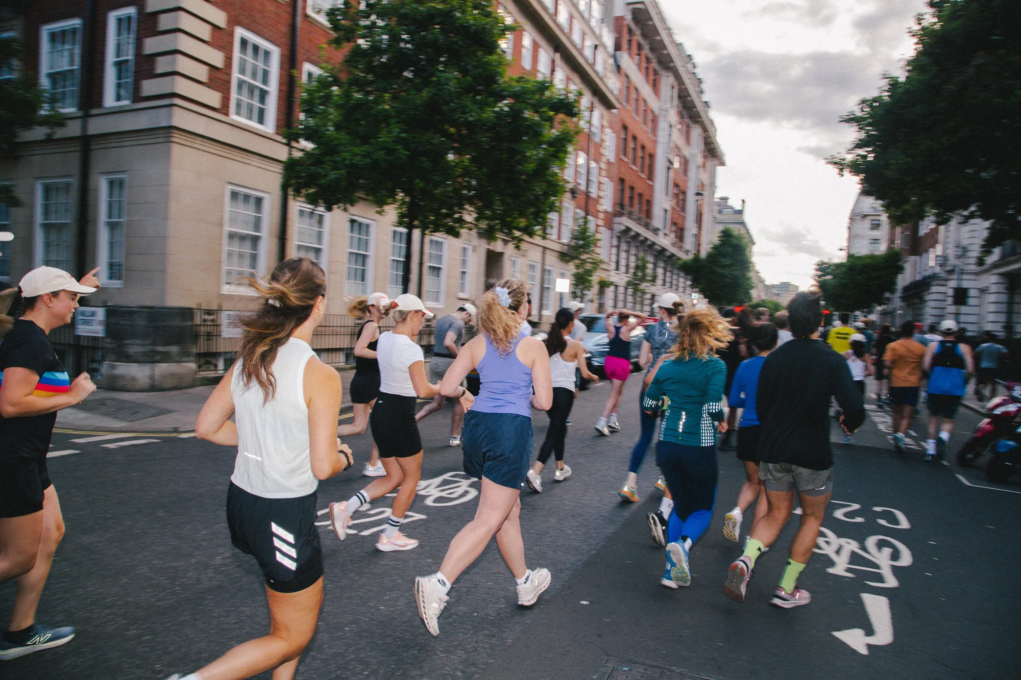 A large group of runners jogs down a city street at dusk, wearing athletic clothing and running shoes. They are running past traditional brick buildings and a designated cycle lane painted on the asphalt.