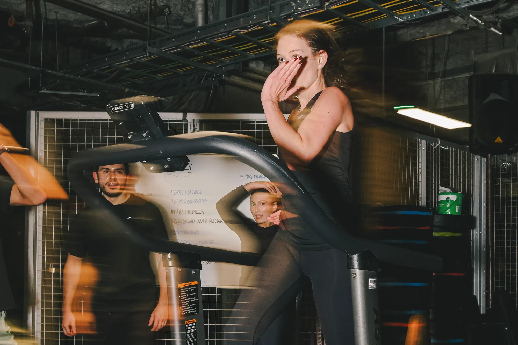 A woman runs intensely on a curved, non-motorised treadmill in an industrial-style gym setting, with motion blur emphasising her speed. A whiteboard listing a challenging workout routine is visible in the background behind the treadmill.