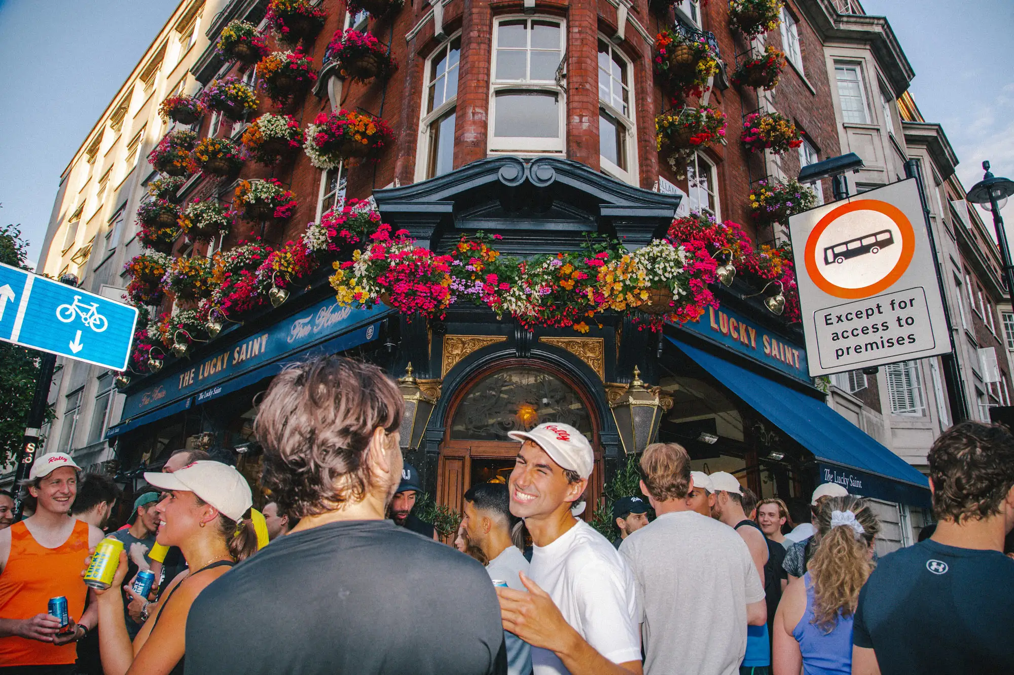 A crowd of people in athletic and casual clothing socialise outside a traditional brick pub named "The Lucky Saint," which is heavily decorated with colourful hanging flower baskets. Several individuals in the foreground are smiling and holding beverage cans.