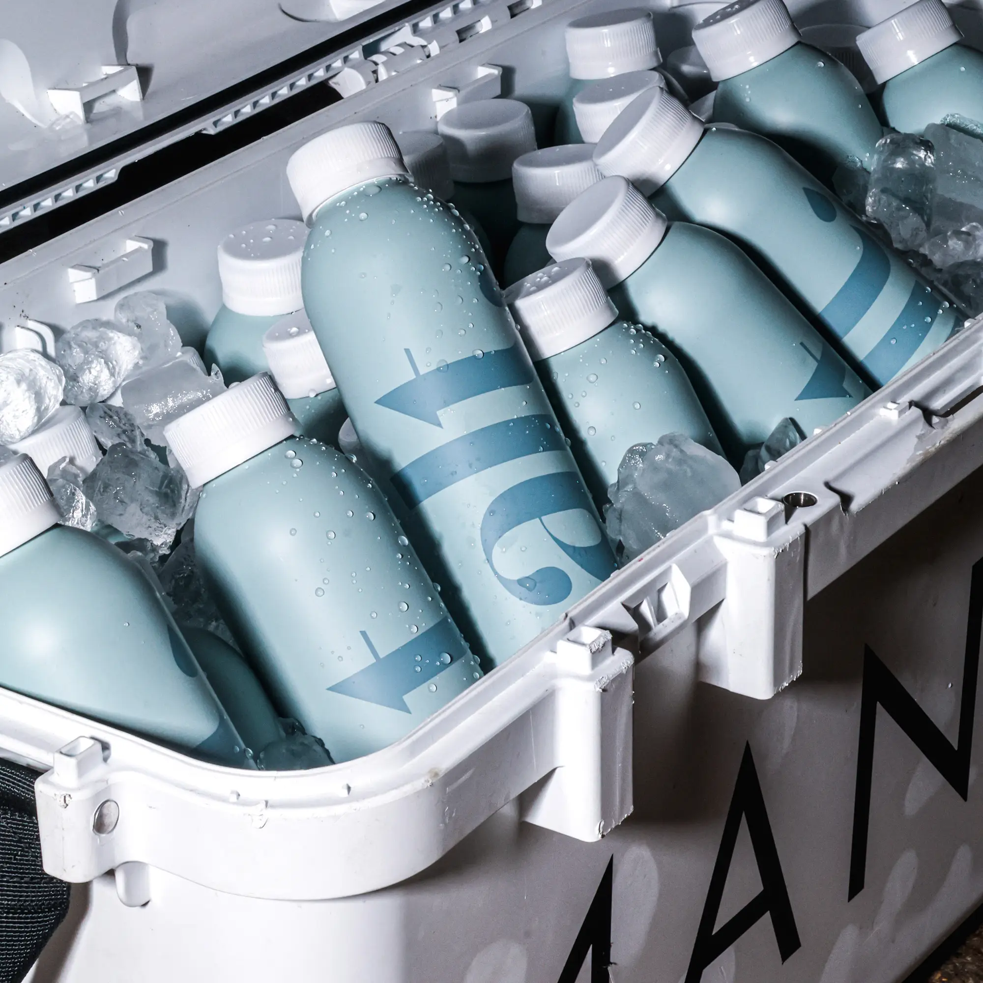 A close-up view of a white cooler filled with crushed ice and several light blue bottles with white caps. The bottles are covered in condensation droplets and feature a darker blue graphic design.