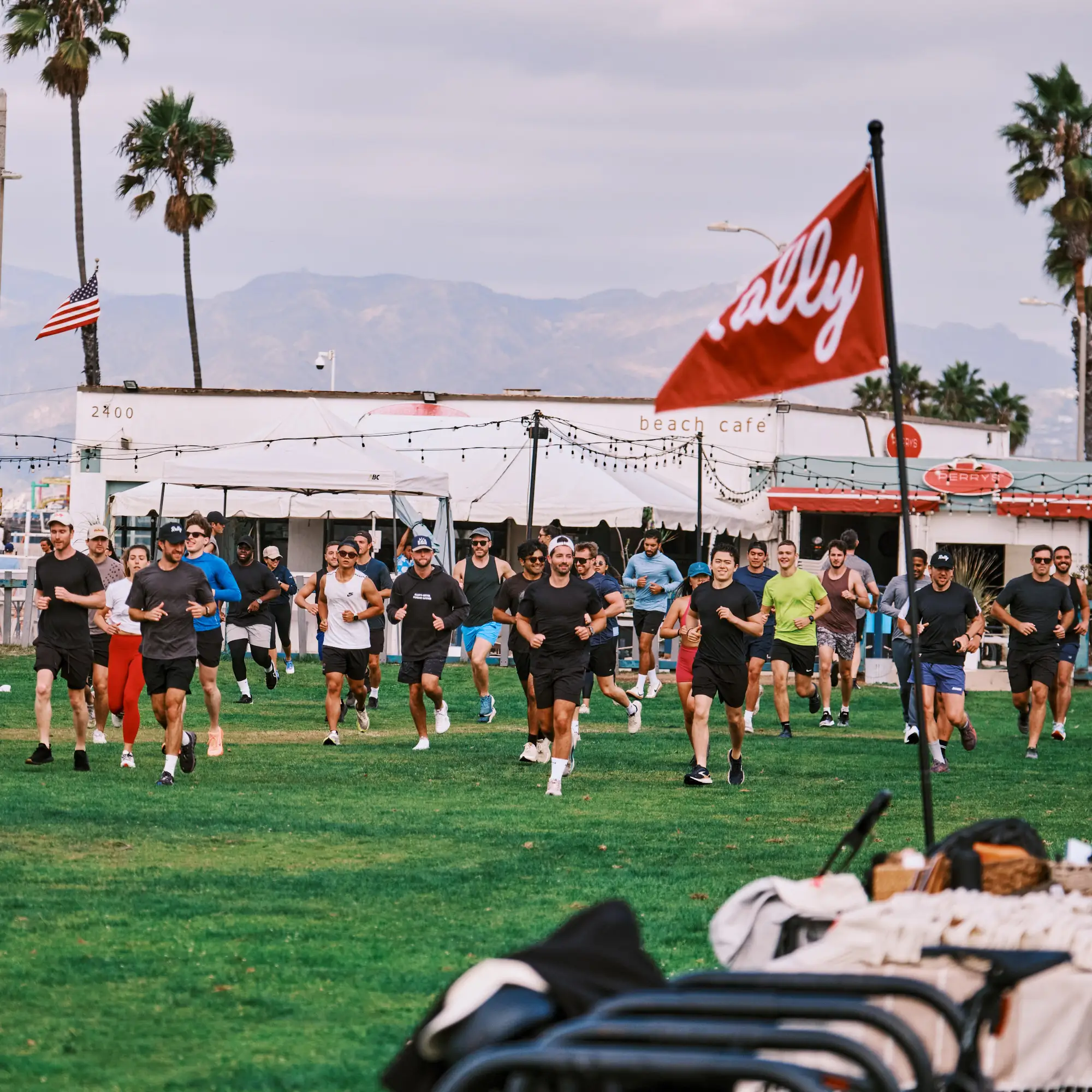 A large group of people running together on a grassy field in front of a white beach cafe. A red flag with the word "Rally" written in white is prominently displayed in the right foreground. Palm trees, a beach area, and mountains are visible in the background under an overcast sky.