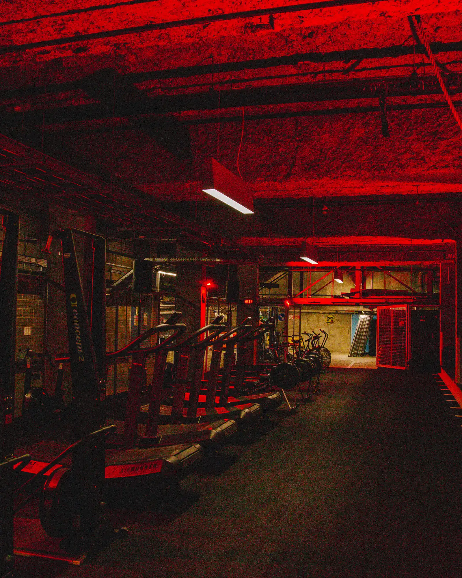 A dimly lit indoor gym or fitness studio illuminated by intense red ambient lighting, featuring an exposed, rough-textured ceiling. A row of curved treadmills and rowing machines line the left side of the room, while a row of stationary air bikes sits further back on the right. A bright rectangular white light fixture hangs from the ceiling, and a doorway with a red metal frame is visible in the background.