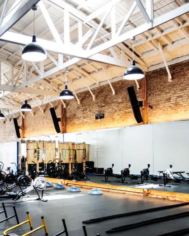 A bright, spacious indoor gym featuring a high, white-painted exposed truss ceiling with skylights and a long exposed brick wall. Several black dome pendant lights hang from the ceiling, and two tall black speakers are mounted on the brick wall. A continuous mirror runs along the lower half of the wall, reflecting wooden equipment storage racks and stationary bikes. The dark gym floor is equipped with rowing machines, blue half-dome balance trainers, heavy battle ropes, and yellow and black agility hurdles.