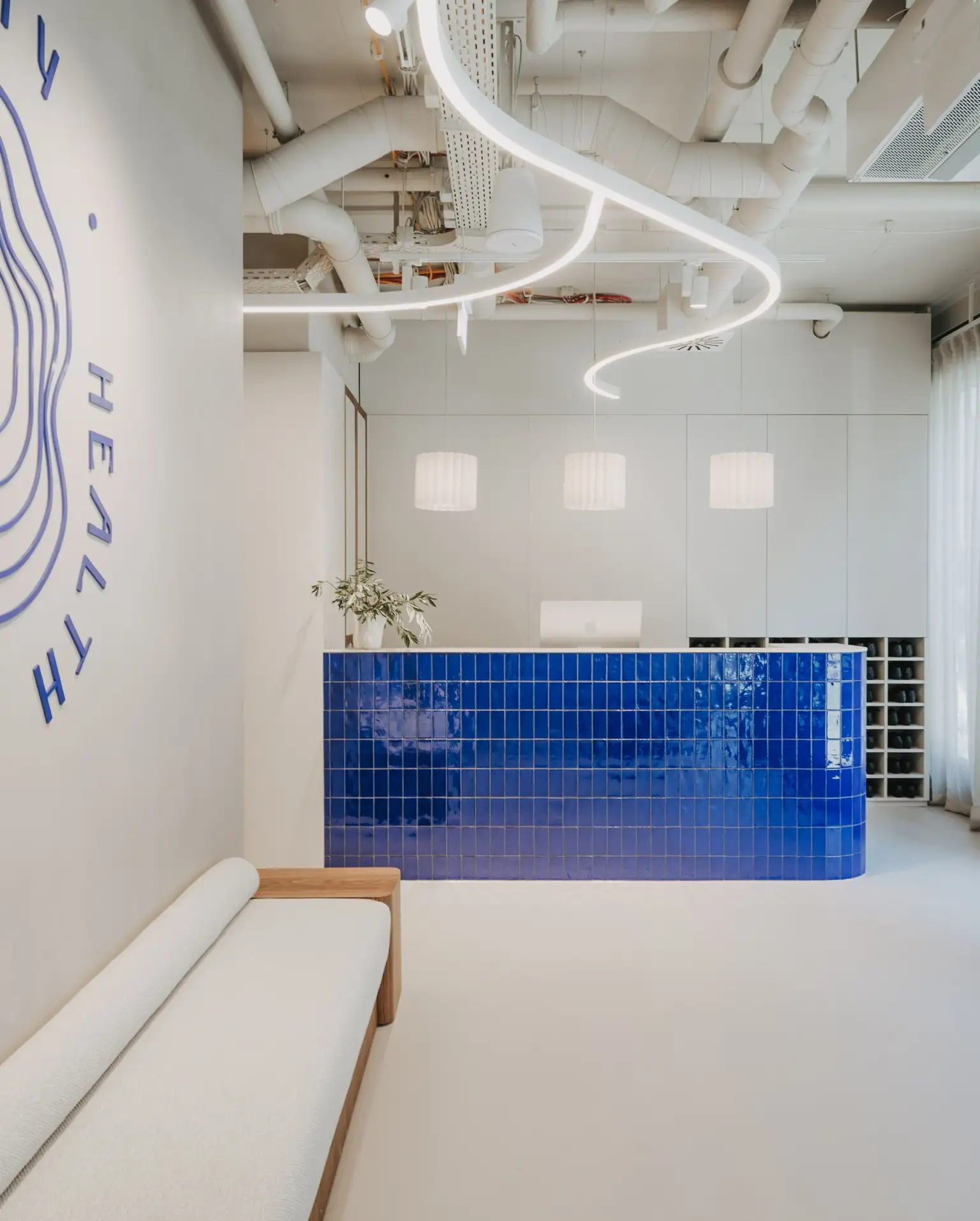 A brightly lit, modern reception area featuring a prominent front desk clad in glossy, cobalt blue square tiles. Three white, fluted pendant lights hang directly above the desk. The ceiling exposes white-painted pipes and ductwork, accented by a striking, continuous winding white LED light fixture. In the foreground on the left, a minimalist white bench with a wooden frame sits against a wall displaying the blue wavy graphics. Behind the reception desk, sleek white cabinetry and a built-in shoe storage cubby complete the space.