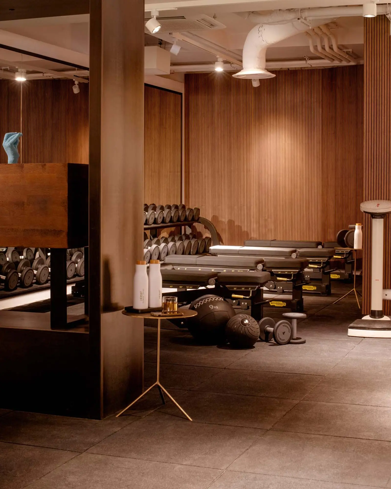An upscale, modern gym interior with a warm ambiance featuring a vertical wood-slat accent wall and dark tiled flooring. Several adjustable workout benches are lined up in the center alongside medicine balls and dumbbells on the floor. In the foreground, a slender gold tripod side table holds two white water bottles with wooden caps and a glass. To the left, a large mirror reflects rows of dumbbells on a storage rack, while the white ceiling features exposed piping and track lighting.