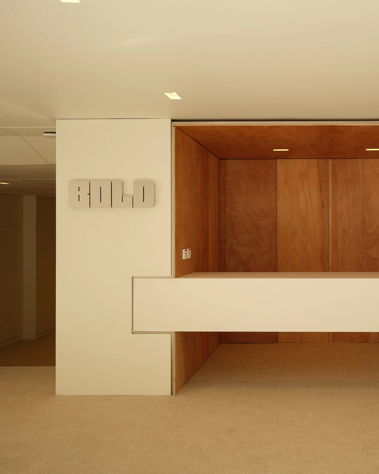 A minimalist reception area featuring a white floating counter against a wood-paneled alcove with the word "BOLD" mounted on the adjacent white wall.