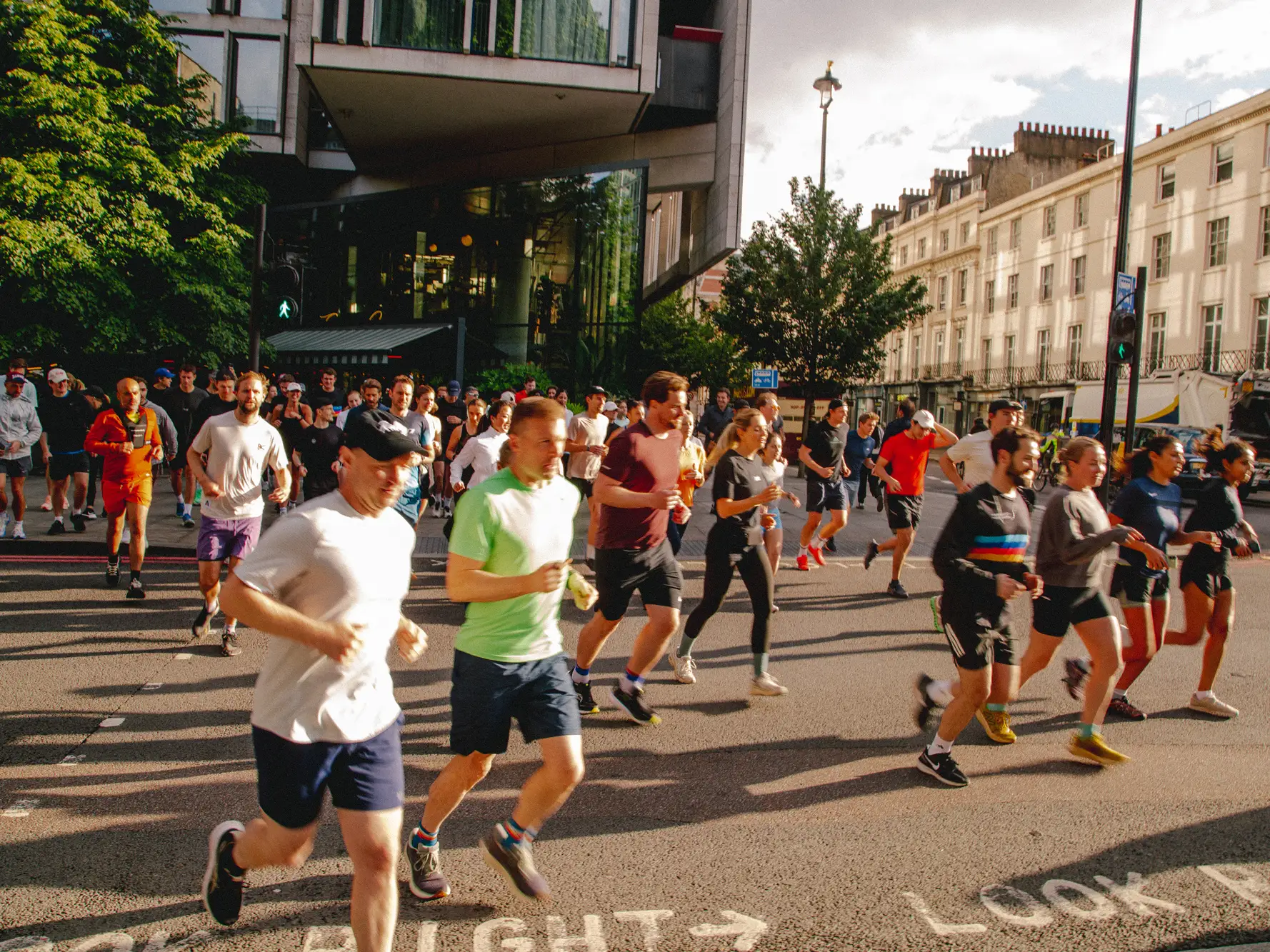 A large group of runners participating in an urban community run, crossing a sunlit city street together.