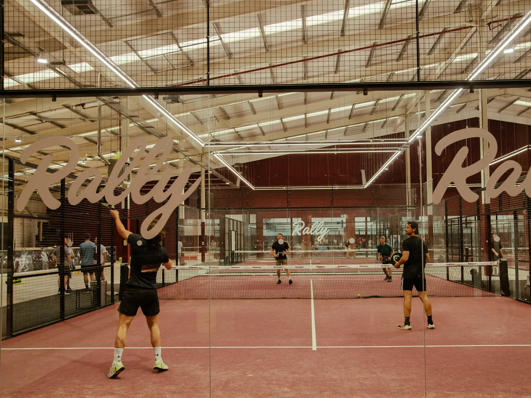 Four people playing a game of padel on a reddish-pink indoor court surrounded by glass walls and mesh fencing under bright linear ceiling lights.